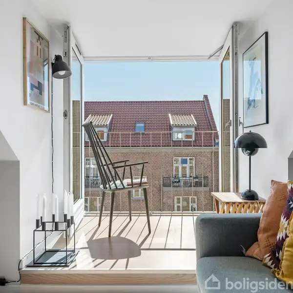 En lænestol står ved åbne altandøre og vender mod en udsigt over bygninger. I stuen er et sofaområde med puder, lamper og indrammede billeder på væggene.