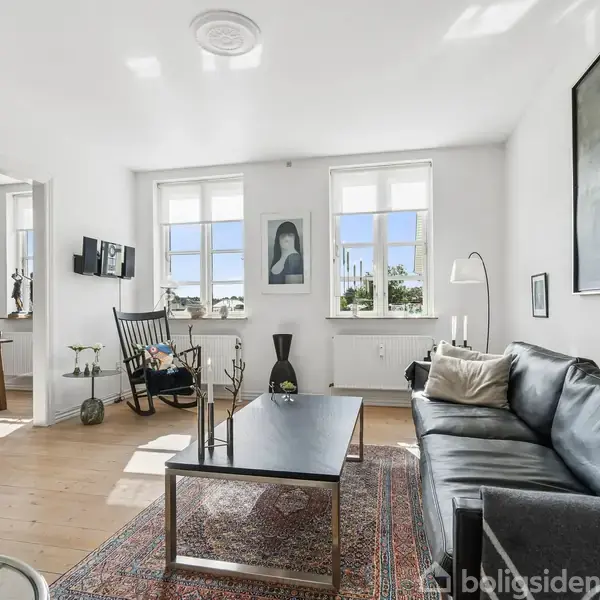 En stue med en sort lædersofa står i et lyst rum med trægulve. Et sofabord er placeret foran en tæppebelagt gulv. Store vinduer lader lys skinne ind, og et maleri hænger på væggen. I baggrunden ses et tilstødende rum med et spisebord og stole.