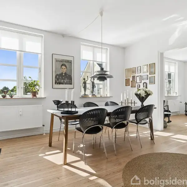 Spisebord med sorte stole står i et lyst rum med trægulv og masser af vægdekorationer. Store vinduer tillader naturligt lys at strømme ind, og en lampe hænger over bordet.