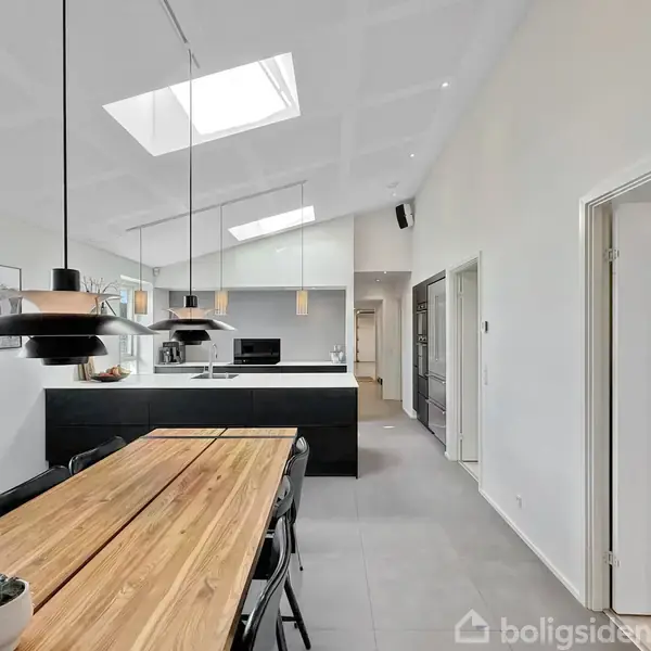 Køkken med langtræsbord i forgrunden, sort køkkenø i midten med hængende lamper over. Lysegrå flisegulv, hvide vægge og ovenlys i loftet skaber et moderne look.