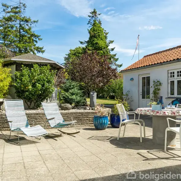 Terrassemøbler står på en solrig, flisebelagt gårdhave. Tre liggestole er placeret nær et bord med stole under en parasol. Baggrunden har grønne træer og et hus med rødt tag.