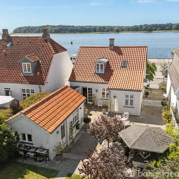 Tre hvide huse med røde tegltage står tæt ved en gårdhave. I baggrunden er en rolig sø med træer på modsat bred. Solrigt vejr præger landskabet.