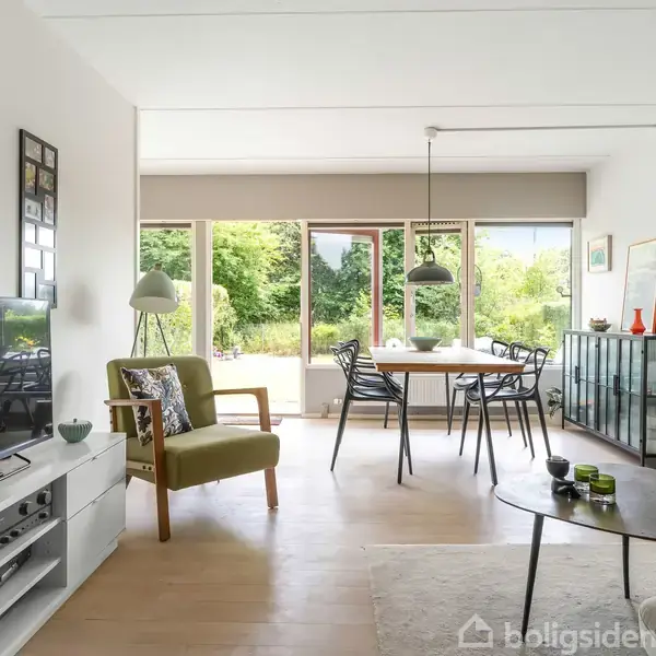 Stue med sofabord og stole, hvor et TV står på et skab. Et spisebord er placeret ved store vinduer, der viser en have. En lampe hænger over bordet.