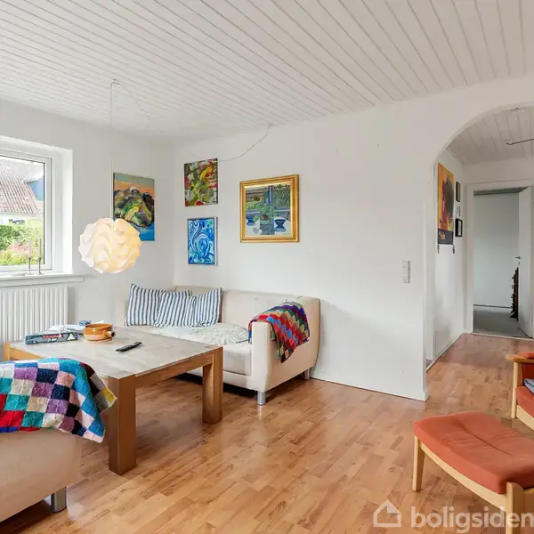 Stue med sofa og lænestole har farverige tæpper, et sofabord og vægkunst. Et vindue på venstre side giver lys, og rummet har trægulv samt en buegang mod en gang.