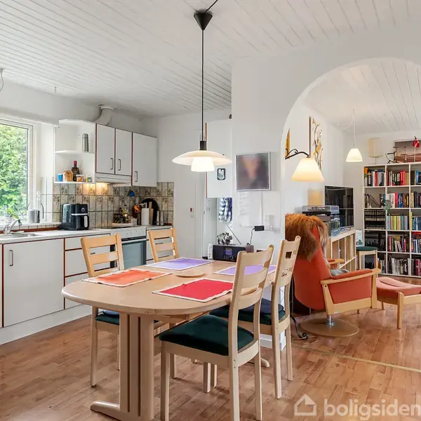 Et lyst køkken-alrum med spisebord og stole, omgivet af køkkenskabe og vinduer med udsigt til grønt. Rummet leder ind til en stue med bogreoler og lænestole.