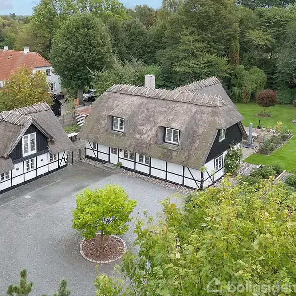 Bindingsværkshus med stråtag ligger i en have med grusbelagt gårdsplads. Omkring er der tæt beplantede buske og træer. Til venstre står en mindre bygning med samme stil.