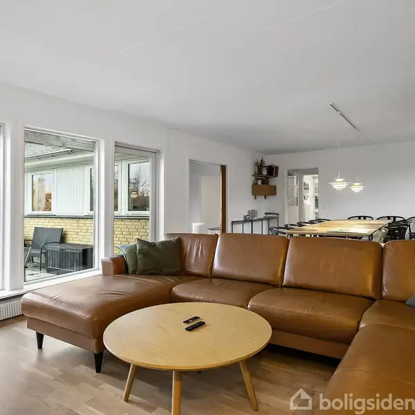 En brun lædersofa står i stuen foran et rundt sofabord. Store vinduer viser en terrasseudsigt. Indendørs er der hvidmalede vægge og træhylder med bøger og genstande.