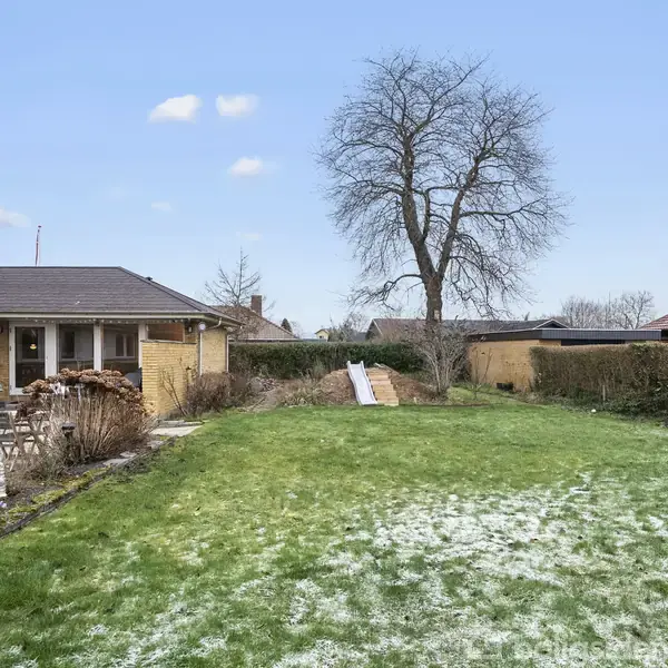 Et hus står ved siden af en stor græsplæne med let sne. I baggrunden ses et stort træ og en hæk. Himmelen er blå med få skyer.