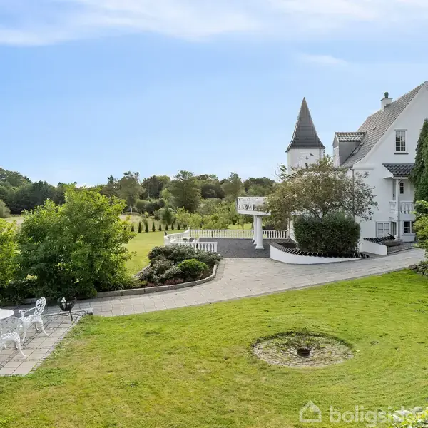 En hvid villa med tårnformet sektion står i en frodig have med terrasse. Til venstre er et drivhus og et havesæt på en flisebelagt overflade. Grønt landskab omgiver sted.