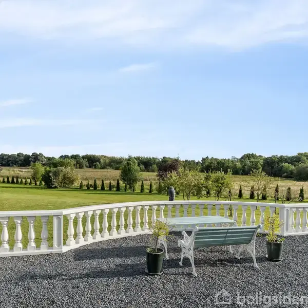 Hvidt havebord med bænke står på en grusbelagt terrasse omgivet af store grønne græsarealer og træer i baggrunden, med en klar blå himmel.