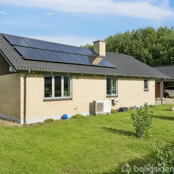 Et enkelt enfamiliehus med solpaneler på taget står på en grøn græsplæne omgivet af buske og træer, under en klar blå himmel.