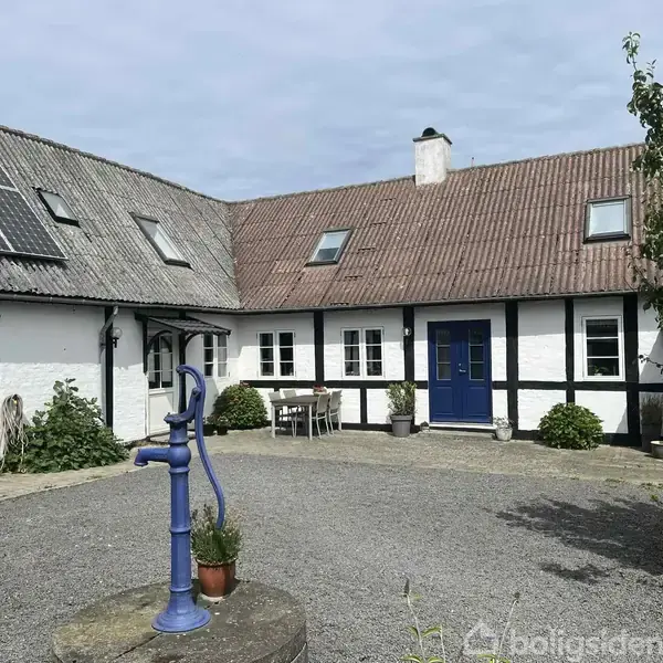 Et hvidmalet bondehus med blå dør og solpaneler på taget omkranser en grusbelagt gårdsplads. En blå vandpumpe står foran huset, omgivet af blomster og buske.