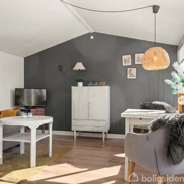 Stue med hvide vægge og en mørkegrå accentvæg. Et tv og skænk står mod væggen. Sofa og stole har pelsbetræk. Rundt sofabord og hængende lampe. Billeder på væggen.