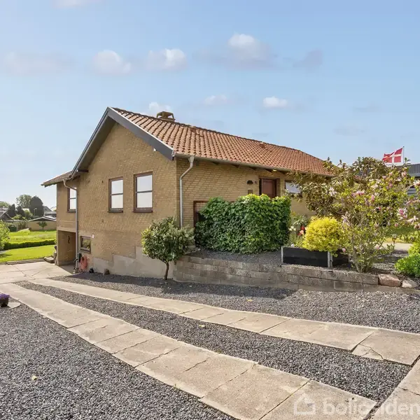 Et gulstenshus står stille omgivet af en velplejet have med buske og flag. Fliser og grus udgør indkørslen, alt under en blå himmel med nogle spredte skyer.
