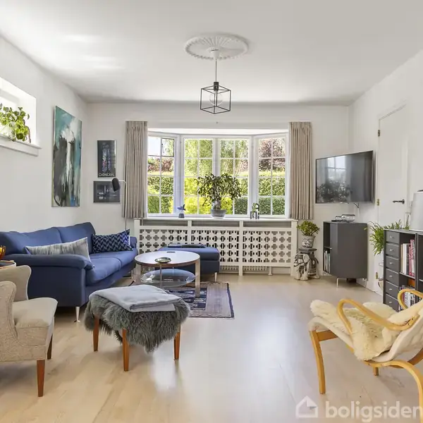 En lys dagligstue med træmøbler indeholder en blå sofa og polstrede stole. Stuen er dekoreret med kunst på væggene og planter ved vinduet, hvilket skaber en hyggelig atmosfære.