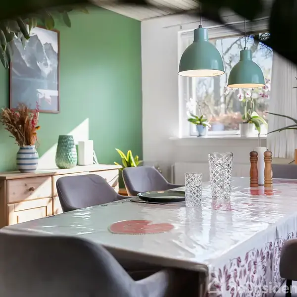 Et spisebord dækket med en plastikdug står i et lyst rum. Der hænger to grønne lamper over bordet. Omkring er der grønne planter, en kommode og vægkunst.