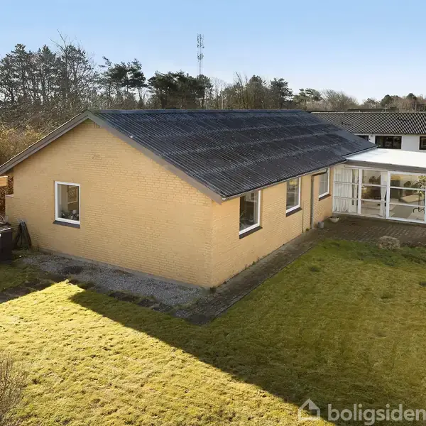 Et gult murstenshus står stille i en have med en vinterhave ved siden. Omgivet af græsplæne og nærliggende huse, er billedet taget under en klar himmel.