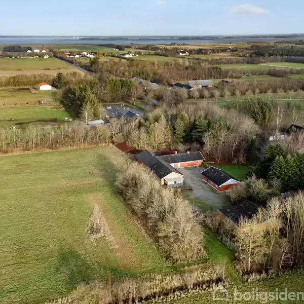 En gård omgivet af træer, beliggende på en stor åben mark med udsigt over spredte landsbyhuse og en fjern horisont.