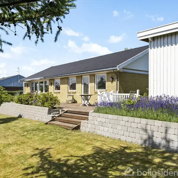 Et enfamiliehus med gule mursten har en terrasse med bord og stole. Det ligger i en have med velholdt græsplæne, lavendel og træer. Himlen er blå med få skyer.