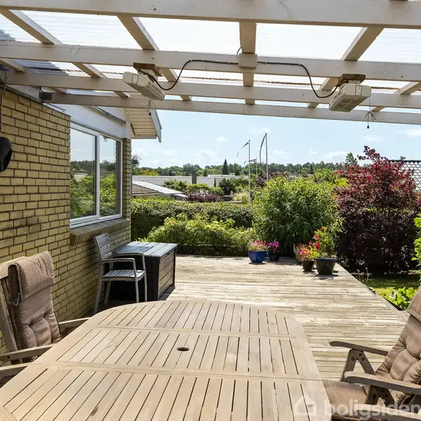 Træbord med stole står på en overdækket terrasse ved siden af et murstenshus. Terrassen rummer også planter i krukker og har udsigt over en grøn have og hustage i baggrunden.