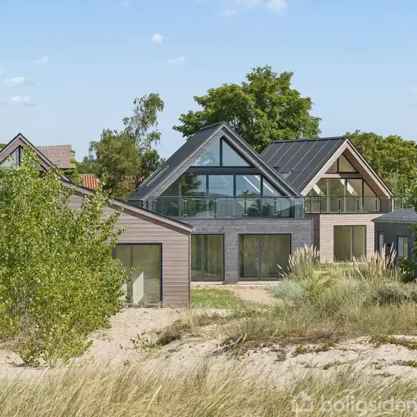 Et moderne hus står stille omgivet af sand og græs ved havet. Huset har store glaspartier og en balkon. I baggrunden ses træer under en klar blå himmel.