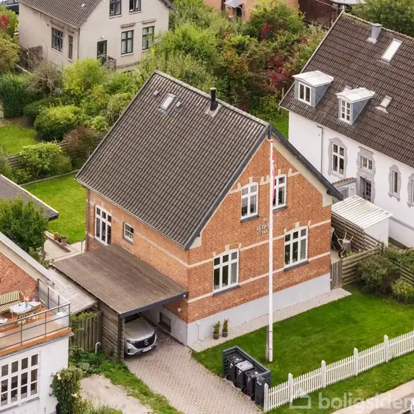 Rødt murstenshus med skråt tag og carport, placeret i en række af lignende huse. Det står i en grøn have, omgivet af andre huse og en hvid stakitport ved indkørslen.