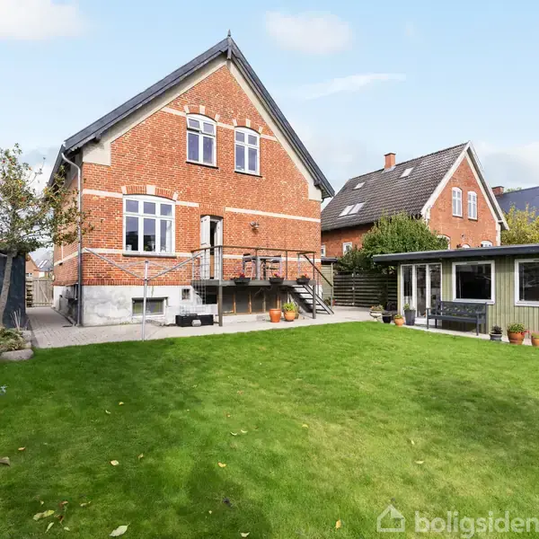 Rødt murstenshus står stille, omgivet af en velholdt grøn græsplæne og flere andre huse. En lille terrasse er forbundet til huset, med en træbygning ved siden af.