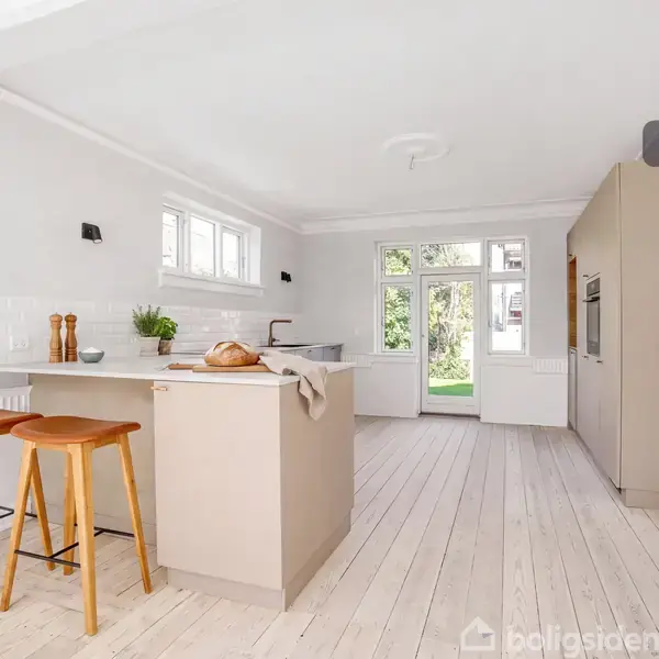 Et lyst køkken med en køkkenø, der har to træstole ved siden af. Køkkenet er moderne med store vinduer, der giver udsigt til en grøn have. Tekst: “Visualisering”.
