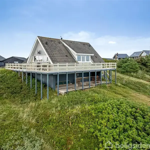 Et sommerhus på pæle står på en græsklædt bakke. Huset har en stor træterrasse og et A-formet tag midt i et grønt område med andre huse i nærheden.