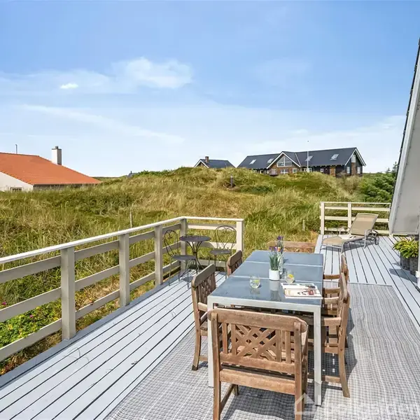 Terrasse med bord og stole står på trædæk foran et moderne hus. I baggrunden ses grønne klitter og huse med røde og sorte tage under en klar, blå himmel.