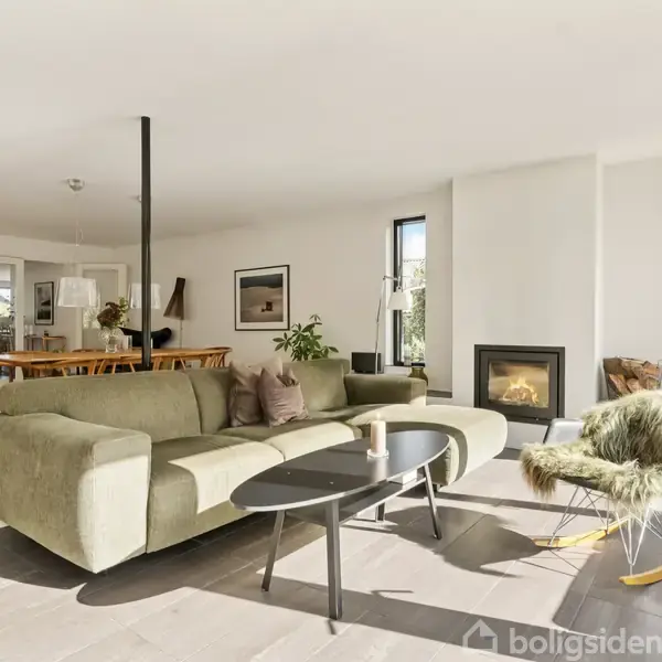 Grøn sofa står i moderne stue med pejs; sofabord foran, gyngestol ved siden af. Spisebord i baggrunden. Store vinduer lader lys strømme ind, skaber lys og luftig atmosfære.