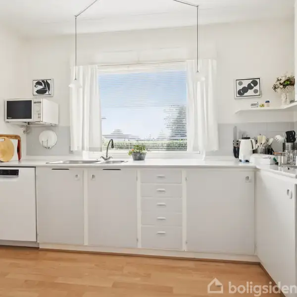 Et lyst køkken med hvide skabe og apparater. Et vindue med gardiner slipper lys ind. Køkkenredskaber og planter er placeret på bordpladen. Trægulv skaber en varm atmosfære.