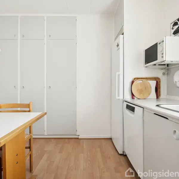 Køkken med hvidt bord og to træstole. Skabe dækker bagvæggen. Mikrobølgeovn er placeret over opvaskemaskinen. Rummet har lyst trægulv og et køkkenvaskområde til højre.