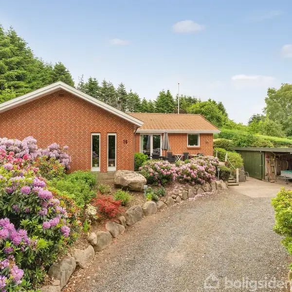 Rødt murstenshus omgivet af blomstrende buske står ved en grusvej. Skovklædt baggrund og carport til højre komplementerer det landlige miljø.