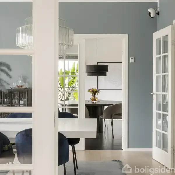 Spisebord med blomsterbuket på, set gennem en åbning mellem to rum. Der er polstrede stole, moderne belysning og blå vægge i en lys og elegant indretning.