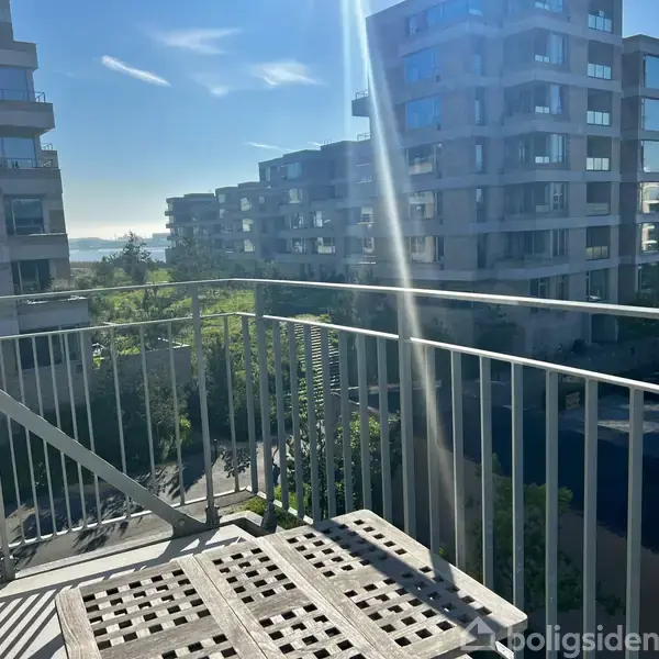 En balkon med en enkel metalgelænder står i sollys. Baggrunden viser moderne lejlighedsbygninger og træer under en klar, blå himmel.