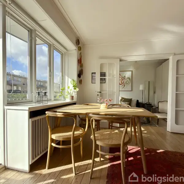 Et spisebord med fire stole står ved siden af et vindue med udsigt til en balkon. I baggrunden ses en stue med åbne dobbeltdøre, lyse vægge og trægulv.