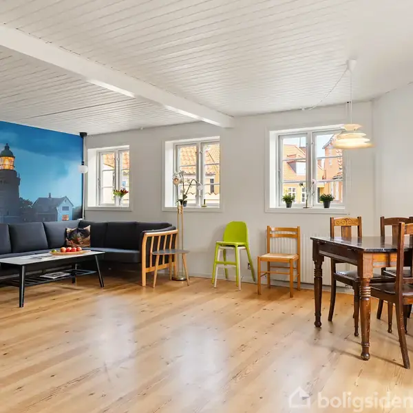 Stue med lys trægulv, hvidmalet loft, og store vinduer. En sofa med et billede af et fyrtårn i baggrunden står til venstre, og spisebord med stole til højre.