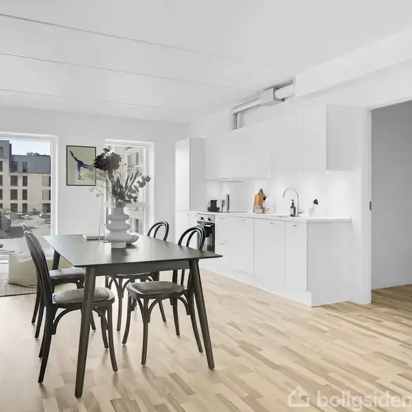 Et spisebord med fire stole står i et lyst køkken. Bordet er pyntet med en vase med blomster. Store vinduer viser en udsigt til moderne bygninger udenfor.