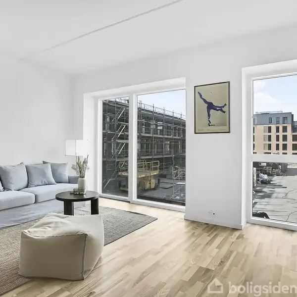 En lys stue med blå sofa står ved siden af store vinduer, som viser et bygningskonstrueret miljø. Et rundt bord og et billede af en akrobat på væggen fuldender rummet.
