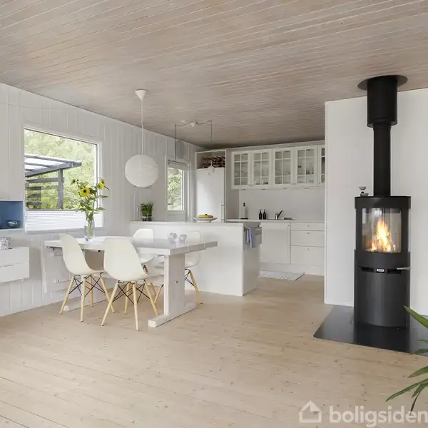 Moderne køkken-alrum med sorte brændeovn, hvide vægge og lofter. Et spisebord med hvide stole står midt i rummet. Indbygget reol og køkkenskabe ses i baggrunden.