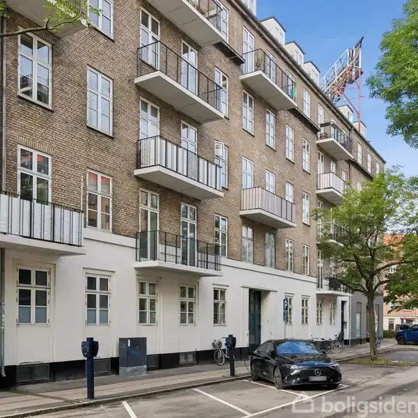 En fem-etagers bygning med murstensfacade og balkoner står langs en gade. Foran er der parkering med en bil, og træer giver skygge. Atmosfæren er bymæssig og rolig.