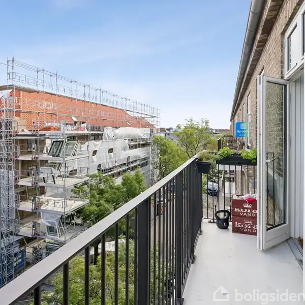 Balkon set fra en lejlighed med åbne franske døre. Byggeplads kan ses i baggrunden, omgivet af stilladser og træer. Klar blå himmel over bymiljøet. Ingen synlig tekst.
