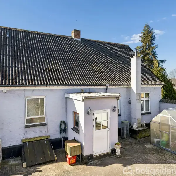 Hvidt hus med sort tag og skorsten; stående i en gårdsplads med en lille tilbygning; omgivet af træer og en bil parkeret ved siden af. En drivhusstruktur ses ved siden af.