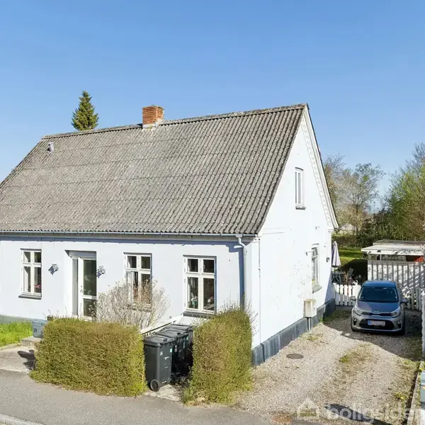 Hvidmalet hus med skråt tag står stille i et beboelseskvarter. En bil er parkeret ved siden af huset. Haven er omkranset af buske og træer.