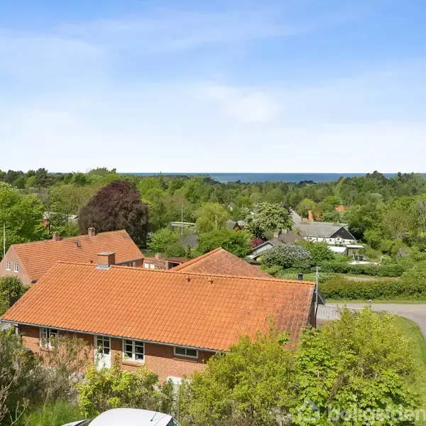 Rødt hus med tegltag omgivet af grønne træer. Beliggenhed i en landlig landsby med udsigt over en vej og havet i horisonten under en klar blå himmel.