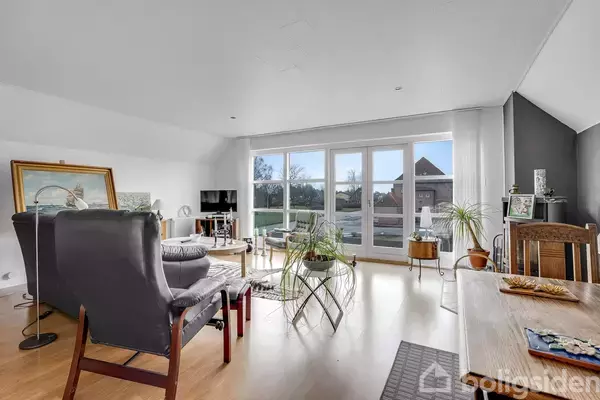 I en lys stue står møbler som lænestole og borde ved store vinduer. Skulpturer og planter tilføjer dekorative elementer. Udenfor ses en have med træer.