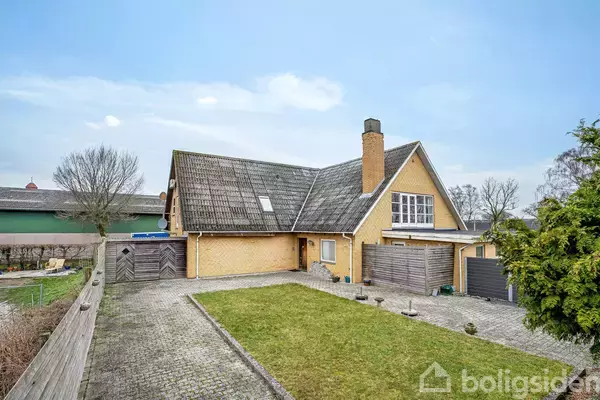 Et gult murstenshus med skråt tag står i en have med flisebelægning og græsplæne, omgivet af træer og en træhegn. Huset har en skorsten og flere vinduer.