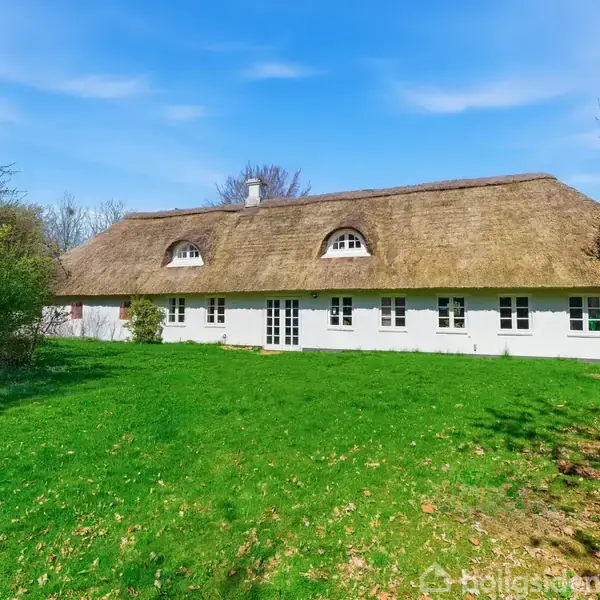 Et stråtækt hus med hvid facade står stille på en grøn græsplæne omgivet af træer under en klar blå himmel.
