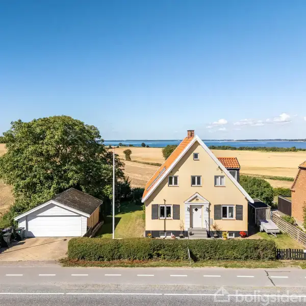 Hus står stille i landligt omgivelser med havudsigt. En garage er til venstre, omgivet af marker og træer under en klar blå himmel.